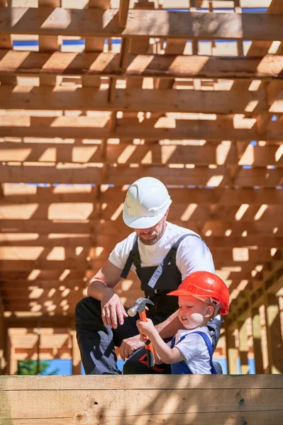Father with toddler son constructing wooden frame house. Son attempting to hammer nail under the guidance of his father on construction site, wearing helmets and overalls. Carpentry and family concept