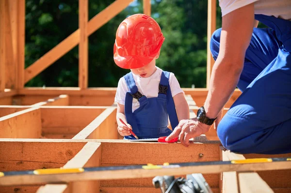 Father with toddler son constructing wooden frame house. Male builder playing with kid on construction site, wearing helmets and blue overalls on sunny day. Carpentry and family concept.