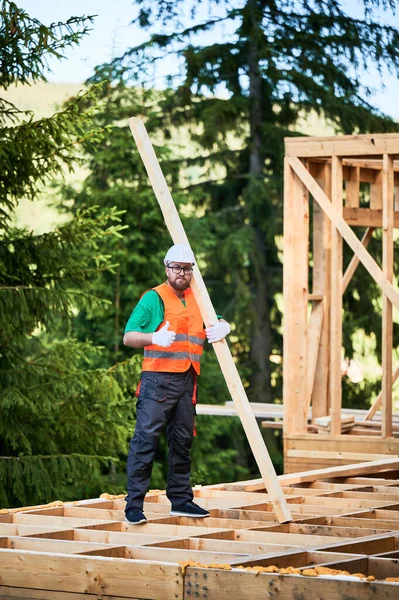 Carpenter builds wooden frame house near the forest. Bearded man with spectacles holds large plank, showing thumbs up, wearing work clothes and helmet. Concept of modern ecological construction.
