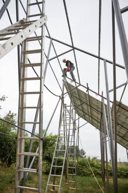 Metal rayların üzerinde duran ve beyaz gökyüzünün altında fotovoltaik güneş paneli sistemi inşa eden güneş sistemi. Erkek işçi güneş modülleri için destek yapısını birleştiriyor.