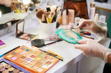 Deri temeli oluşturan steril eldivenli kadın ellerini kapat. Makyajcı kadın makyaj masasının yanında göz farı paleti ile duruyor ve profesyonel makyaj için temel hazırlıyor..