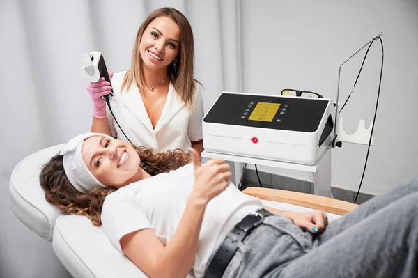 快乐美容师和年轻女性患者经激光皮肤护理后表现出认可的姿态 美容师持用激光扫描仪进行皮肤再生处理 — 图库照片