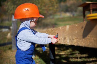 Boy toddler playing as builder on construction site. Child carpenter in orange helmet and blue overalls learning to build wooden frame house outdoor on sunny day. Carpentry and workshop concept.