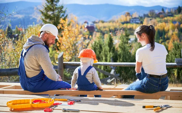 Back view of father, mother and son building wooden frame house. Toddler boy, daddy and woman having a rest on construction site. Guys wearing helmet and blue overalls. Carpentry, family concept.