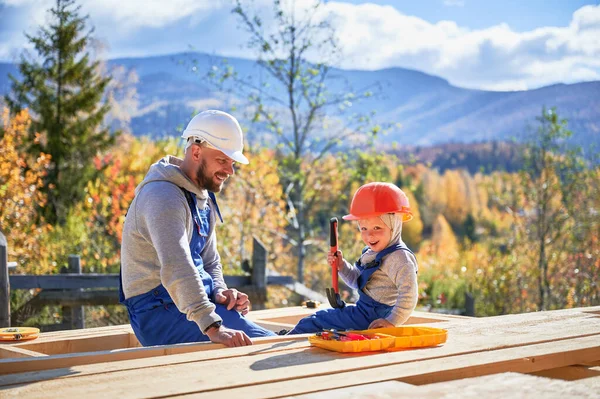 Father with toddler son building wooden frame house. Male builders hammering nail into plank on construction site, wearing helmet and blue overalls on sunny day. Carpentry and family concept.