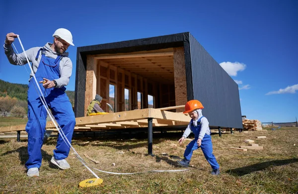 Father with toddler son building wooden frame house in the Scandinavian style barnhouse. Male builder and kid playing with tape measure on construction site on sunny day. Carpentry and family concept.