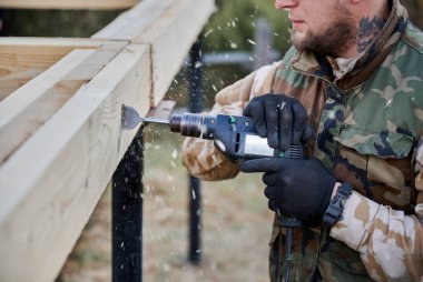 Yığınla temele ev inşa eden bir adam. İnşaat alanındaki müstakbel evin ahşap çerçevesinde elektrikli matkapla delik açan erkek işçiyi kapatın. Marangozluk kavramı.