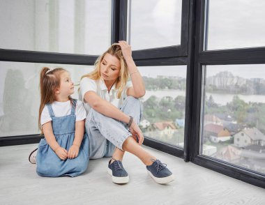 Bir kadın ve küçük bir kız yerde oturuyor ve yeni bir aile evinde birbirlerine bakıyorlar. Anne ve çocuk yeni inşa edilen apartmanın panoramik penceresinden manzaranın keyfini çıkarıyorlar.