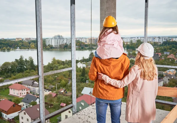 父母带着女儿站在在建公寓楼内的背景图 男人和女人带着孩子戴着建筑用的头盔 从未来的家欣赏城市美景 — 图库照片