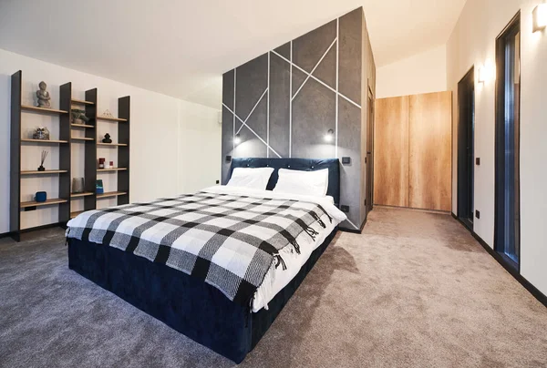 stock image Simply furnished sleeping area with big bed and wooden shelves with buddhist decoration at home. Side view of dark bed covered with plaid bedspread against gray wall in bedroom. Concept of interior.