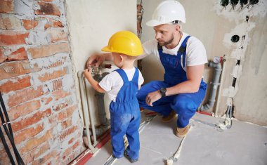 Baba ve küçük çocuk tadilat sırasında apartmana boru tesisatı döşüyorlar. Erkek tesisatçı ve çocuk, güvenlik miğferi takmış. Evde tadilat yaparken musluğu kontrol ediyorlar..