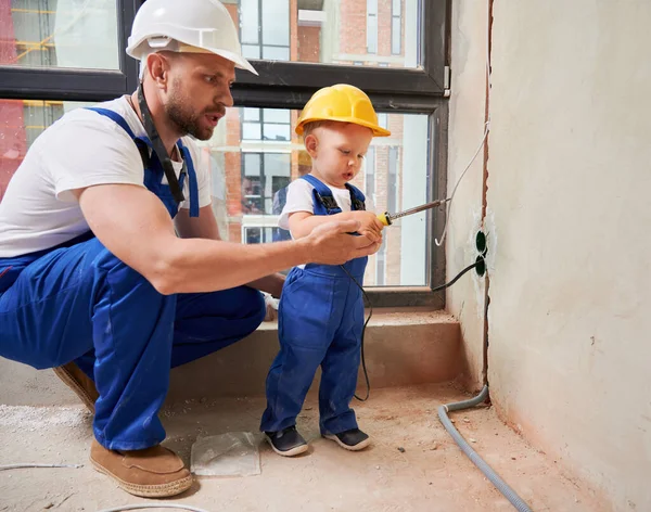 Erkek işçi ve küçük çocuk, daireye elektrik kabloları ve prizler yerleştirirken kaynak onarım aletlerini kullanıyorlar. Elektirikli lehimleme demiri olan bir adam ve çocuk ev onarımı üzerinde çalışıyor..