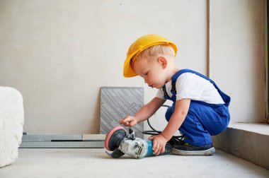 Elektrikli öğütme makinesi tutan küçük sevimli inşaat işçisinin yan görüntüsü. Beyaz duvara yaslanırken iş tulumu ve güvenlik kaskı takan sevimli bir çocuk..