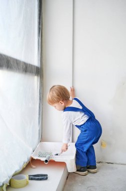 Küçük inşaat işçisi çocuk pencerenin yanında duruyor ve dairede beyaz duvar boyuyor. Çalışan bir çocuk evde tamirat sırasında boya fırçası kullanıyor..