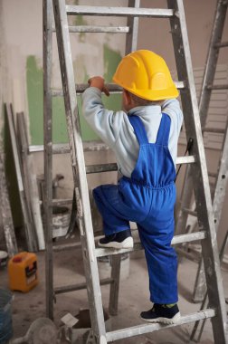 Ev tadilatında çalışırken çocuk inşaat işçilerinin merdivene tırmanma görüntüsü. Güvenlik kaskı takan ve iş tulumlu bir çocuk apartmanda merdivenlerden çıkıp tadilat yaptırıyor..