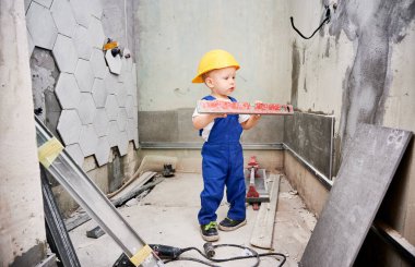 Çocuk inşaat işçisi ruh seviyesinde bir alet tutuyor. Kasklı ve iş tulumlu bir çocuk apartman banyosunda duruyor. Ev yenileme ve çocukluk konsepti.