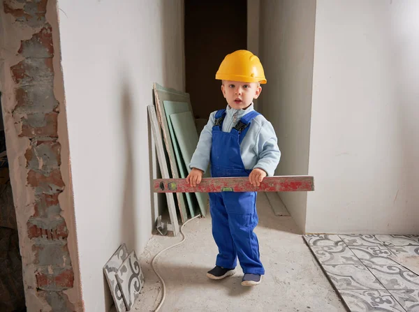 Çocuk inşaat işçisi, elinde ruh seviyesinde bir aletle kameraya bakıyor. Güvenlik kaskı takan ve iş tulumu giyen bir çocuk evin duvarında duruyor, tadilatta..