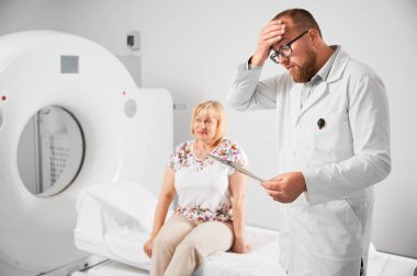 Tıbbi bilgisayarlı tomografi ya da MRI tarayıcısı. Şok olmuş, şaşırmış doktor tomografiye bakıyor, MRI sonuçlarını inceliyor. Arka planı bulanık bir şekilde koltukta oturan bir kadın hasta. Sağlık hizmetleri kavramı.