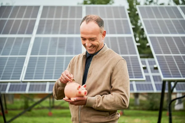 stock image Male putting his money in piggy bank for future profit. Young man making investment in alternative ecological resources. Man in casual clothes holding moneybox on background of solar panel.