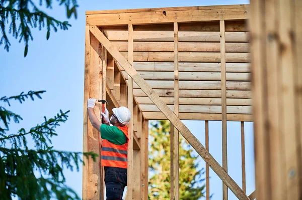 stock image Carpenter constructing wooden frame house near the forest. Bearded man hammering nails into structure while wearing protective helmet and construction vest. Concept of modern ecological construction.