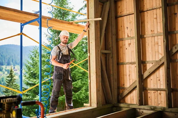 Marangoz ormanın yakınında iki katlı ahşap çerçeve inşa ediyor. Çekiç tutan sakallı bir adamın portresi. Koruyucu kask ve tulum giymiş. Ekolojik modern yapı kavramı.