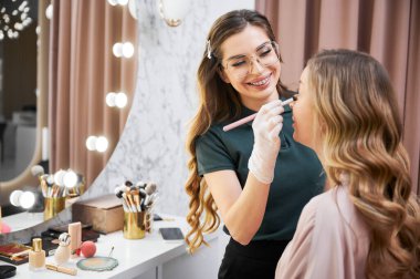 Gözlüklü, gülümseyen kadın stilist. Makyaj fırçasıyla müşterinin yüzüne fondöten uyguluyor. Neşeli güzellik uzmanı, yüz makyajı stüdyosunda kadın için profesyonel makyaj yapıyor.