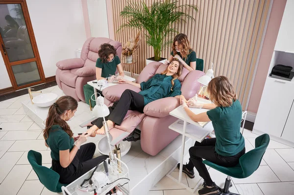 Woman relaxing in comfortable salon chair while having manicure, pedicure and eyelash extension procedure. Manicurist, pedicurist and eyelash specialist working with client in modern beauty salon.