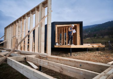 Male inspector building wooden frame house on pile foundation. Man builder standing on construction site in safety helmet, with construction documentation inspecting quality of work.
