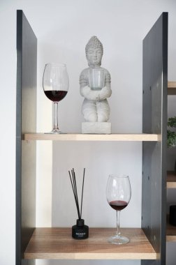 İçinde Buda heykeli, tütsü çubukları ve şarap kadehleri olan ahşap bir raf. Evdeki beyaz duvara karşı biblo ve aroma çubuklarıyla dizayn edilmiş küçük rafın yakın görüntüsü. Dekor kavramı.
