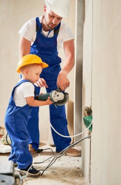 Kablo kanalları ve prizleri babasıyla birlikte duvara yerleştirirken elinde elektrikli tel değirmeni tutan çocuk. Güvenlik miğferi takmış bir adam ve çocuk elektrikli aletler kullanıyor ve evdeki elektrik kablolarını takıyor..
