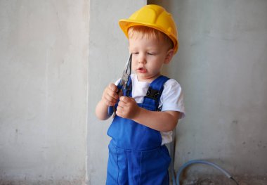 Adorable baby boy looking at instrument in building under construction. Cute child wearing work overalls and safety helmet while playing with tool at construction site.