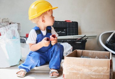 Yerde karton kutunun yanında oturan sevimli küçük çocuk. Evini yenilemek için gerekli aletler ve malzemelerle. Çocuk inşaat işçisi apartmanda inşaat malzemesi tutuyor..