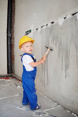 Çekiç tutan ve duvarın yanında kameraya bakan bir oğlan çocuğu. Çocuk güvenlik kaskı takıyor ve tadilat sırasında evde inşaat malzemesi kullanırken tulum giyiyor..