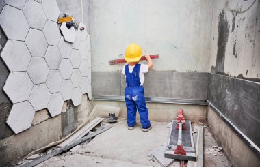 Küçük çocuk dairede ruh seviyesi olan duvar yüzeyini kontrol ediyor. İş tulumları ve güvenlik miğferi takan çocukların ev tadilatında kullandıkları düzgün aletlerin arka görüntüsü.