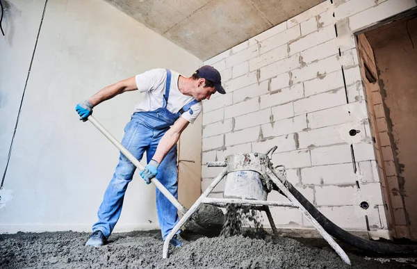 Male worker using shovel while shoveling sand-cement mix in building under construction. Man preparing floor screed material while standing near concrete screed mixer machine.