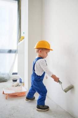 Evin duvarını boyayan küçük çocuk tadilat altında. Güvenlik kaskı takan ve evde tamirat sırasında boya silindiri kullanan çocuk..
