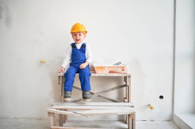 Cute kid construction worker sitting on wooden table against white wall in apartment under renovation. Cheerful little boy wearing safety helmet and work overalls while playing at home.