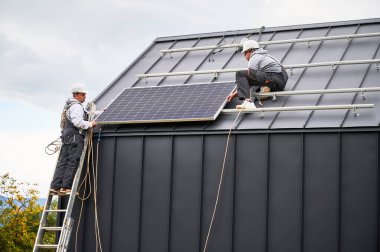 Çatıya fotovoltaik güneş modülü istasyonu inşa eden atçılar. Miğferli elektrikçiler güneş paneli sistemi kuruyor. Alternatif ve yenilenebilir enerji kavramı.