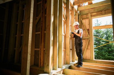 Marangoz ahşap çerçeveli ev inşa ediyor. Tornavidayla çalışan, iş tulumu ve kask giyen bir adam. Modern çevre dostu yapı kavramı.