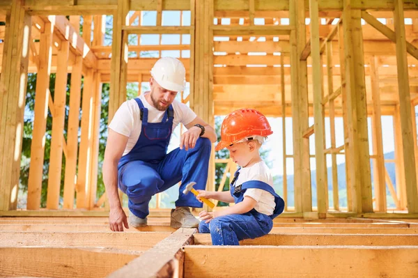 Father with toddler son building wooden frame house. Worker instructing his son on art of hammering nails on construction site, wearing helmets and overalls. Carpentry and family concept