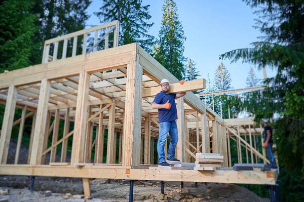 Carpenter builds wooden frame house near the forest. Bearded man holds large plank on the shoulder, showing thumbs up, wearing work clothes and helmet. Concept of modern ecological construction.