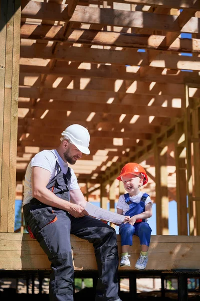 Father with toddler son building wooden frame house. Guys having fun on the edge of balcony, examining the construction plan, wearing helmets and overalls on sunny day. Carpentry and family concept.