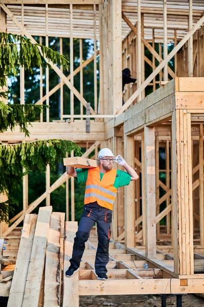 Carpenter building wooden-framed house near the forest. Man holding a large beam on his shoulder, dressed in work clothes and helmet. Idea of modern and sustainable construction.