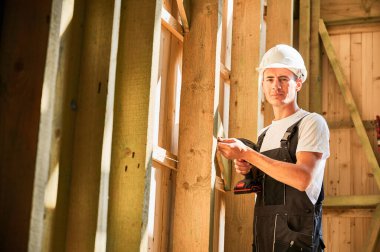 Marangoz ahşap çerçeveli ev inşa ediyor. Tornavidayla çalışan, iş tulumu ve kask giyen bir adam. Modern çevre dostu yapı kavramı.