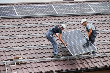 Evin çatısında fotovoltaik güneş modülü taşıyan erkek işçiler. Kasklı elektrikçiler dışarıda güneş paneli sistemi kuruyorlar. Alternatif ve yenilenebilir enerji kavramı.