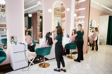 Genç kadınlar aynaların önünde otururken kuaförler müşterilere kuaförlük yapıyor saç ve makyaj sanatçıları profesyonel makyaj yapıyor. Modern güzellik salonunda çalışan makyaj uzmanı ve kuaför..