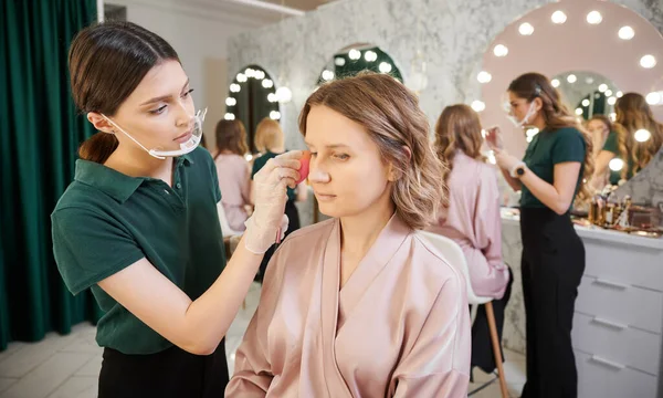 Female beauty specialist applying foundation on woman face with blending sponge. Woman sitting at dressing table while makeup artist doing professional makeup in protective face mask.