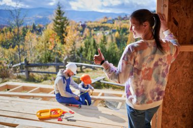 Father, mother and son building wooden frame house. Toddler boy helping his daddy, while woman looking for them on construction site. Guys wearing helmet and blue overalls. Carpentry, family concept.