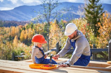 Küçük oğlu olan baba ahşap çerçeve inşa ediyor. Erkek müteahhitler, güneşli bir günde kask ve mavi tulum giyerek inşaat alanında tahtaya çivi çakıyorlar. Marangozluk ve aile kavramı.