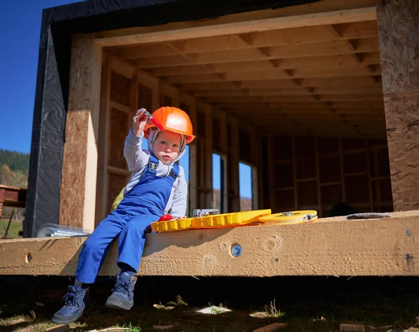 Boy toddler playing as builder on construction site. Child carpenter in orange helmet and blue overalls learning to build wooden frame house outdoor on sunny day. Carpentry and workshop concept.